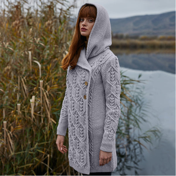 Woman wearing a gray knitted coat standing by a body of water with mountains in the background.