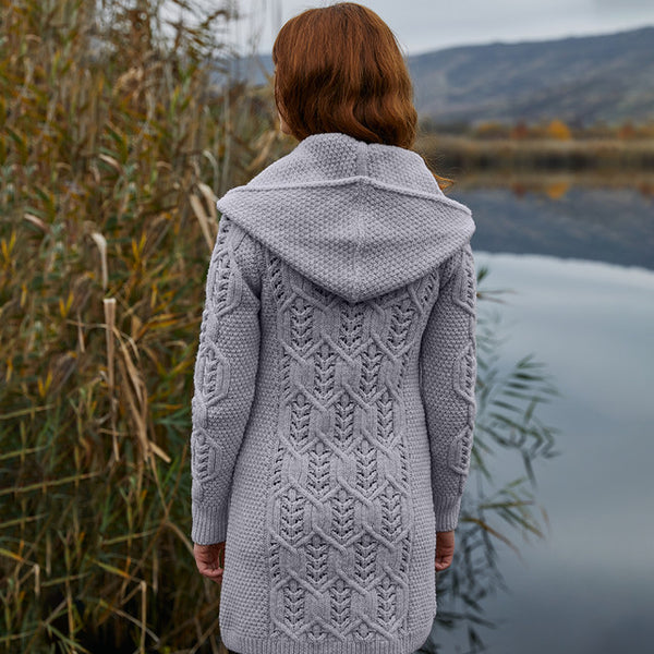 Person wearing a gray knitted hoodie standing by a body of water with mountains in the background.