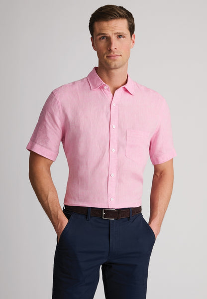 A man wearing a light pink linen short-sleeve button-down shirt and navy blue trousers, standing against a neutral gray studio background.