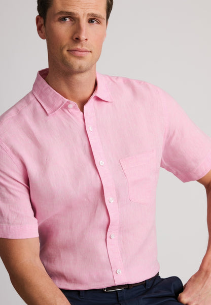 A man wearing a light pink linen short-sleeve button-down shirt and navy blue trousers, sitting in a neutral gray studio background.